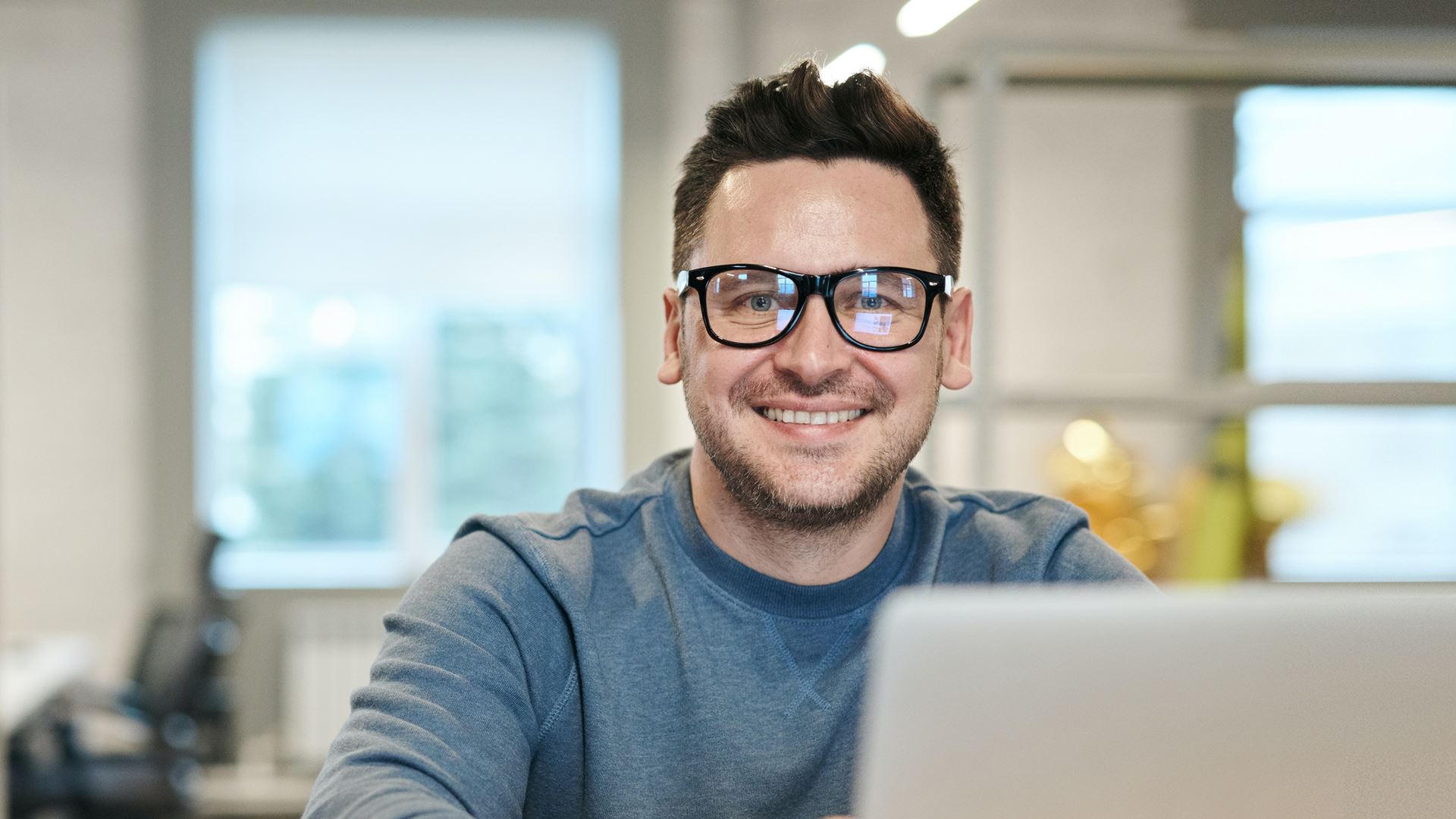 Man met bril achter laptop lachend in camera kijkend